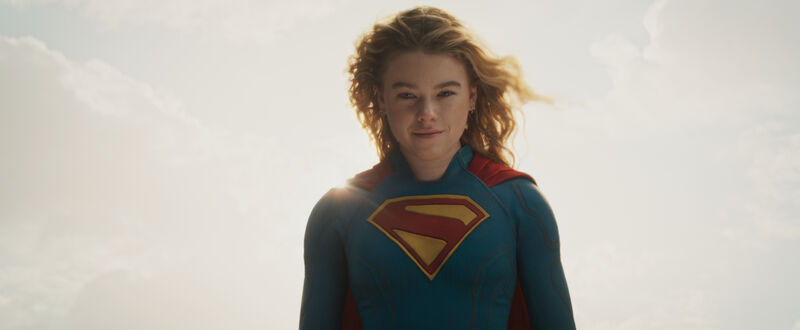 A costumed superhero stands against a bright sky, wearing a blue suit with a red cape and a large emblem on the chest.