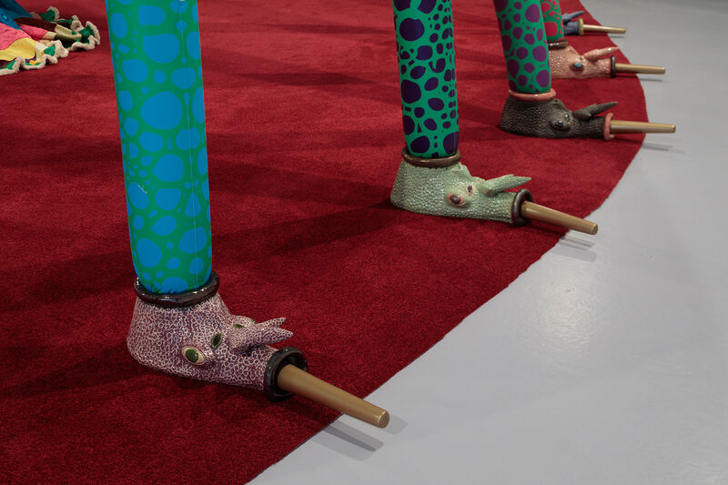 A close up of ceramic elements of an insect-like structure which stands on a red rug. The ceramics have faces and long noses, and are glazed in earthy pastel tones.