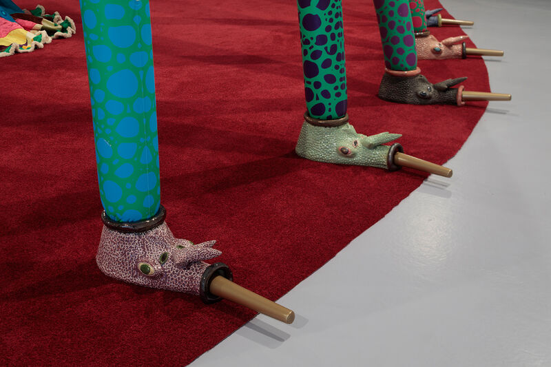 A close up of ceramic elements of an insect-like structure which stands on a red rug. The ceramics have faces and long noses, and are glazed in earthy pastel tones.