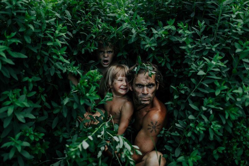 A man crouches in a bush with two small children, they are posing in a mock-fierce way.