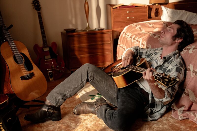 A man sits on the floor with his head on the bed, playing guitar
