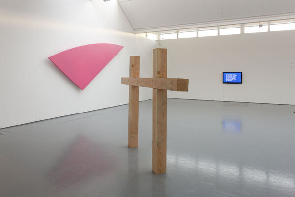 From Jonathan Horowitz's exhibition. A large pink shape, with two straight side and one curved, hangs on the gallery wall. In the middle of the room, there are two plain wood crosses which are touching one another.