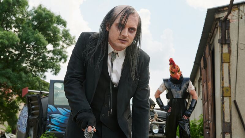 Elijah Wood as a man in a suit with long, thinning hair. A person in a chicken mask stands in the background