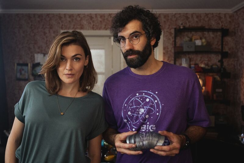 A man and woman look nervously at the camera. He holds a metal slinky.