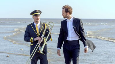 A man in a suit and a man in a marching band uniform holding a trombone stand on a beach.