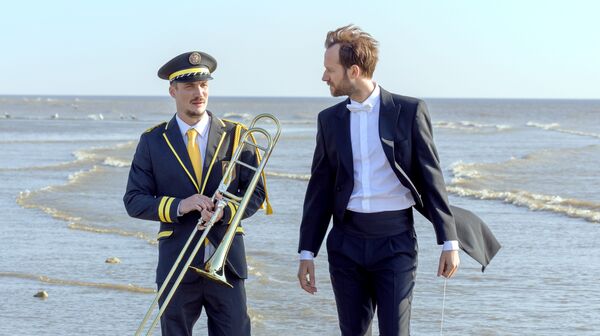 A man in a suit and a man in a marching band uniform holding a trombone stand on a beach.