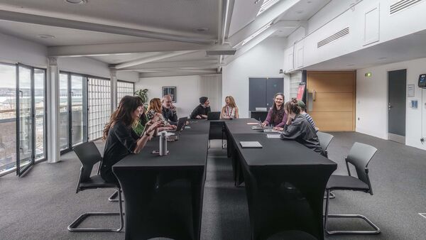 A room in an office with people sitting around a u shaped table having discussions big windows in the background