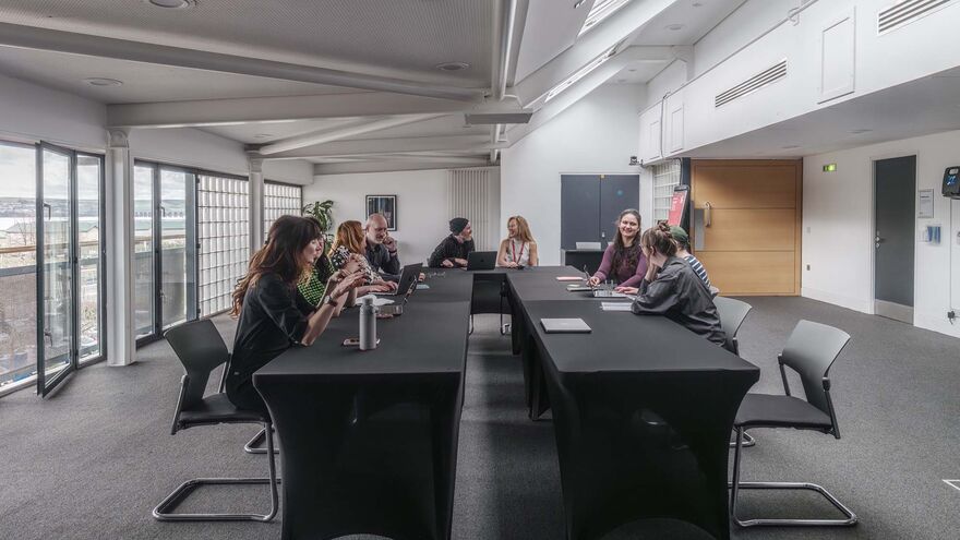 A room in an office with people sitting around a u shaped table having discussions big windows in the background