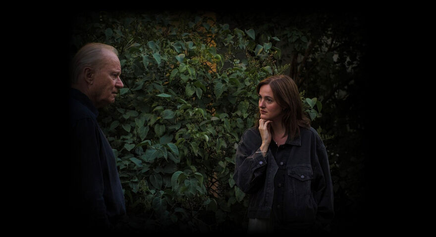 An older man and a young woman stand next to each other by a hedge.