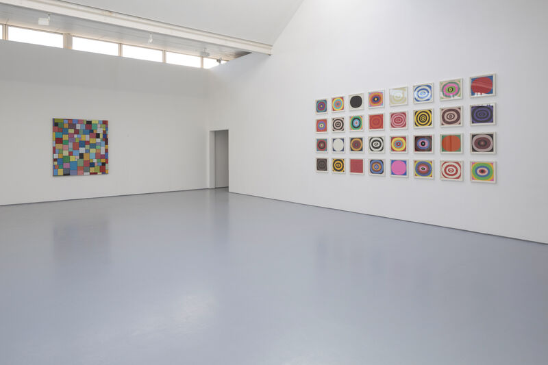 Image shows a brightly lit gallery with natural light, white walls and grey floor. On the walls to the left and right are colourful 2d artworks. To the right, these are small and arranged in a grid. To the left there is one work composed of many small squares.
