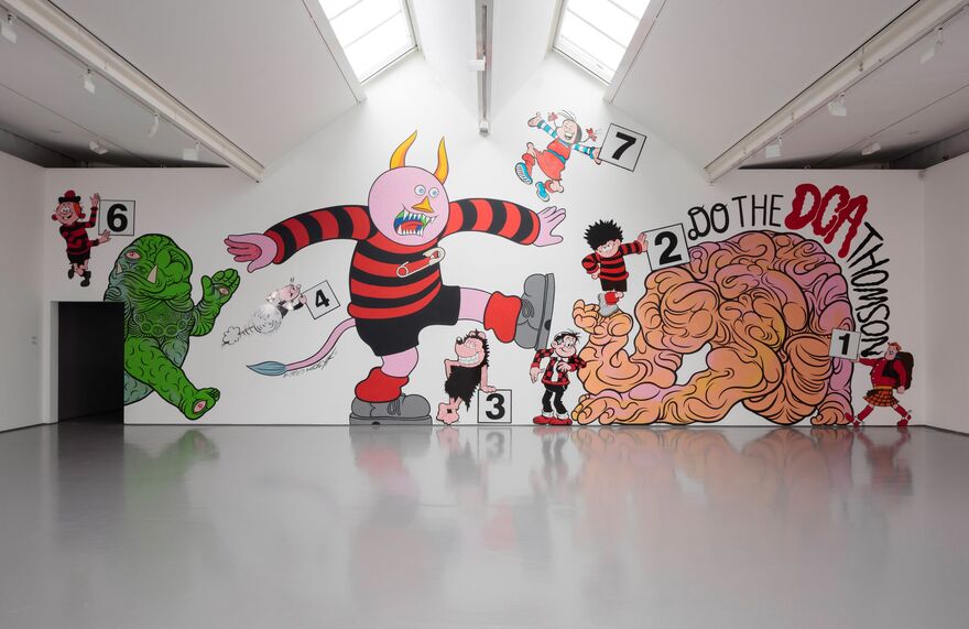 On the white gallery wall in DCA, a giant mural shows a horned creature wearing a Dennis the Menace jumper, surrounded by other DC Thomson characters holding numbered signs, and a strange green monster. Writing on the wall says 'Do the DCA Thomson'