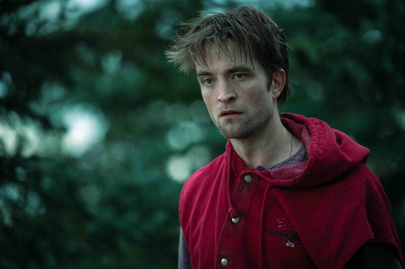 A bedraggled man in an ill-fitting red hoodie stands outside