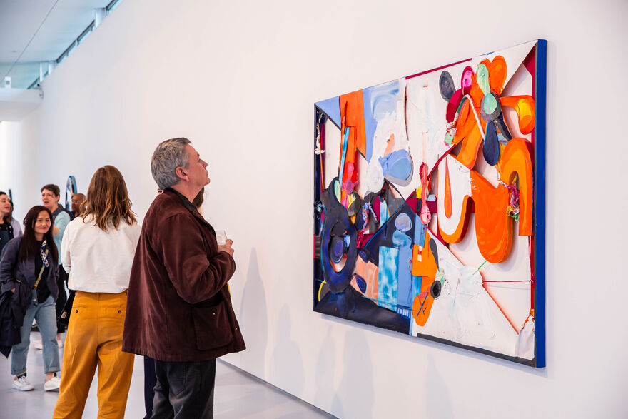 A man looks at a colourful, textural painting hung on the white wall of a gallery. 