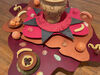 A close up of a sculptural work with a vase, colourful cut out shapes layered on top of each other, ceramic bowls and fruits. The objects are painted in earthy warm colours and stacked with the vase on top. 
