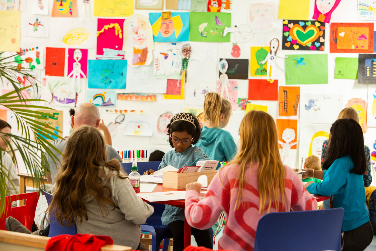 Children sitting around tables in Gallery 1 doing art making. The wall behind them is covered in children's artwork