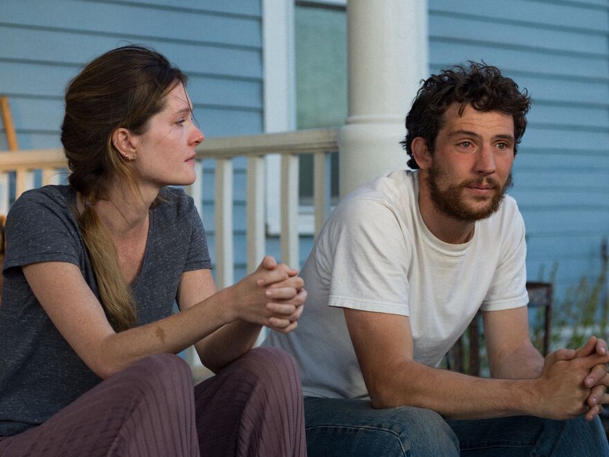 A man and woman sit side-by-side looking emotional with tears in their eyes outside a blue trailer home
