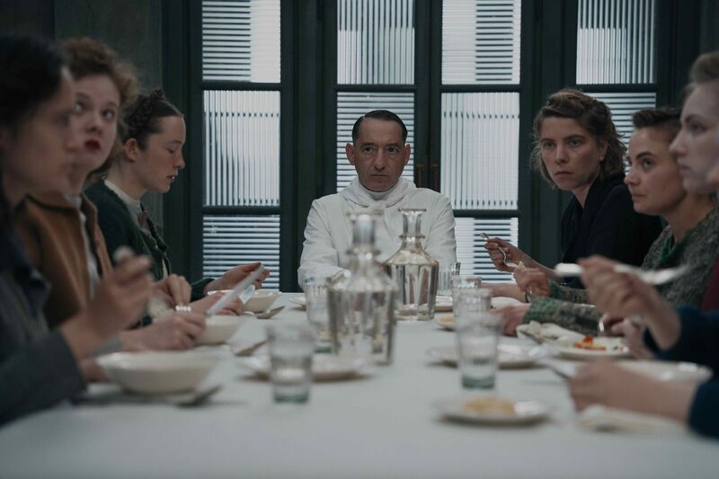 A group of 1940s women, and a man dressed all in white, sit at a dining table