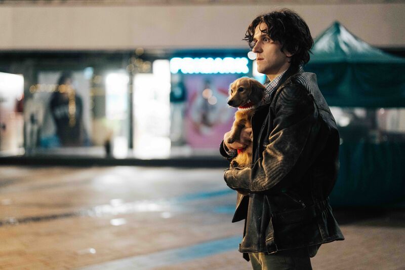 A man with floppy dark hair stands in the street holding a daschund.