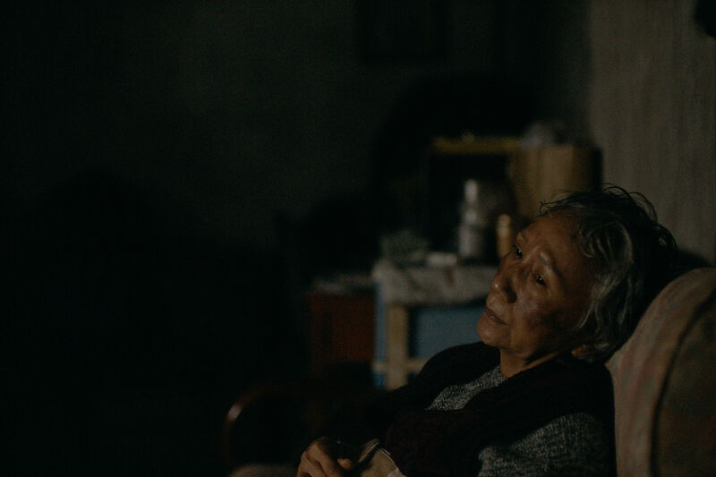 An older woman sits in a dark room with a neutral expression