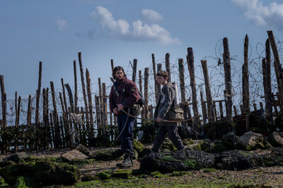 A man and a young boy walk along a fence made of trees and barbed wire, carrying crossbows.