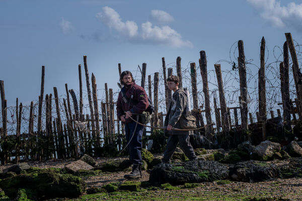 A man and a young boy walk along a fence made of trees and barbed wire, carrying crossbows.