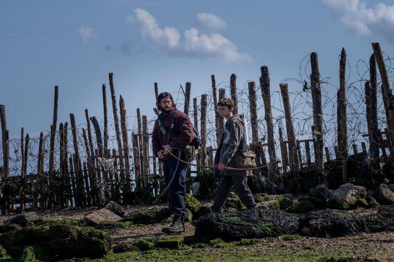 A man and a young boy walk along a fence made of trees and barbed wire, carrying crossbows.