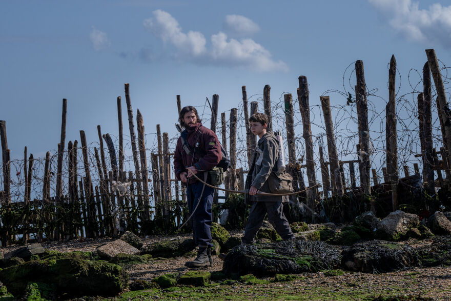 A man and a young boy walk along a fence made of trees and barbed wire, carrying crossbows.