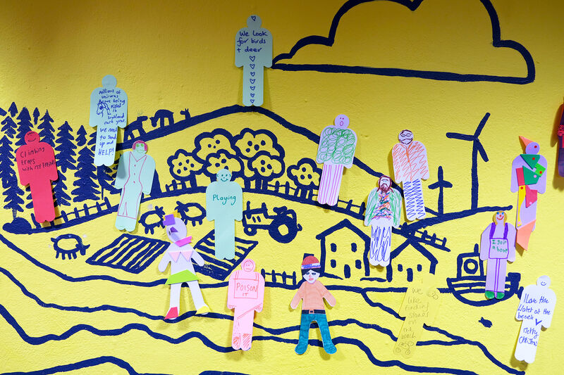 Yellow wall painted with a landscape with paper people cut out and places on top