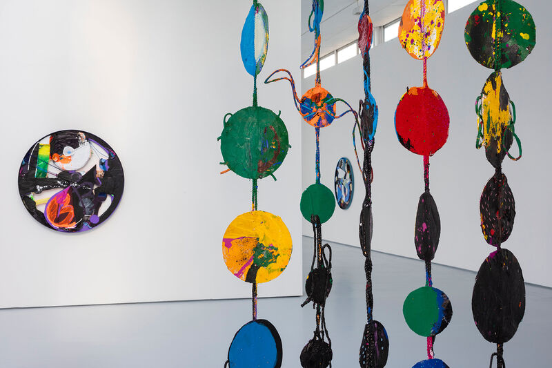 A close up of an artwork hanging from the ceiling, this image shows the colourful found materials and braiding that constitute a hammock-like structure. In the background a round canvas mainly using black and pink paints can be seen on a white partition wall.  