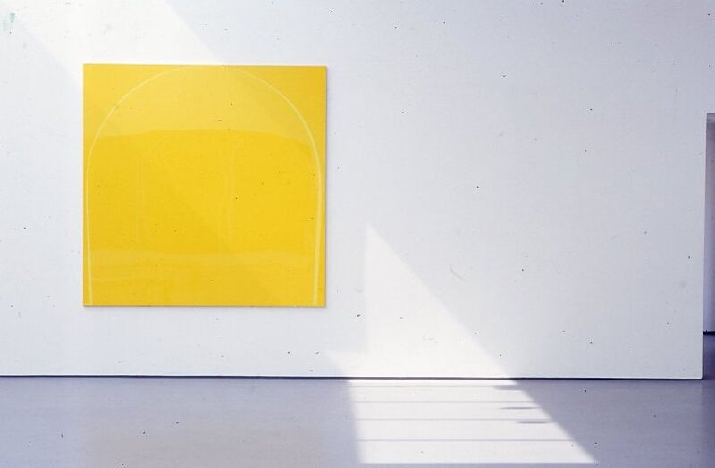 A bright yellow square painting hangs on a. white wall.