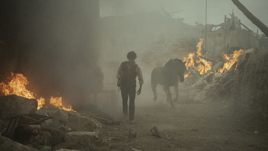 A man walks through burning rubble while a horse runs behind him
