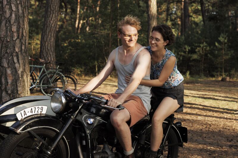 A man and woman in 1940s clothing ride on a motorbike, smiling.
