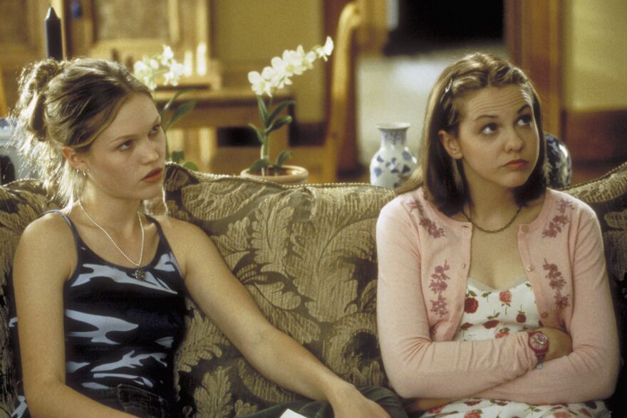 A still from 10 Things I Hate About You. Two teenage girls who look grumpy sit on a couch.
