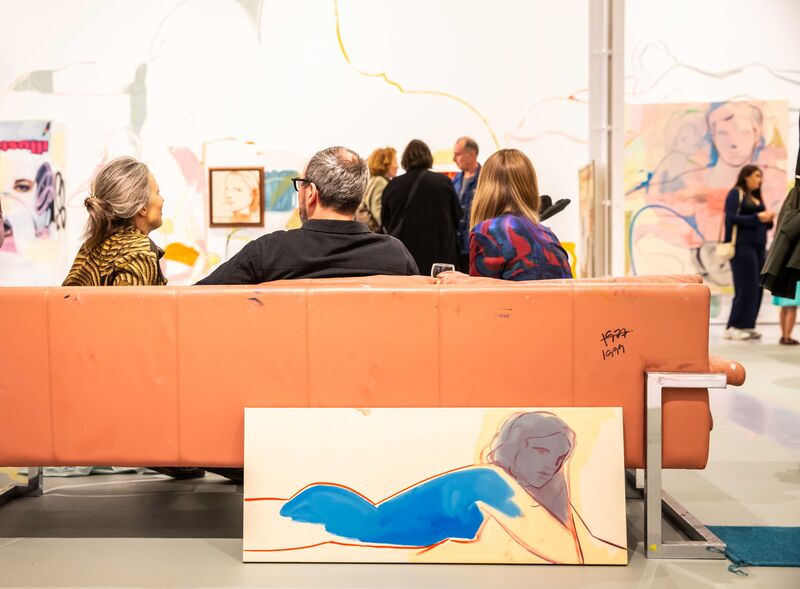 People sit in a gallery space on an orange leather sofa wit paintings on the wall in the background 
