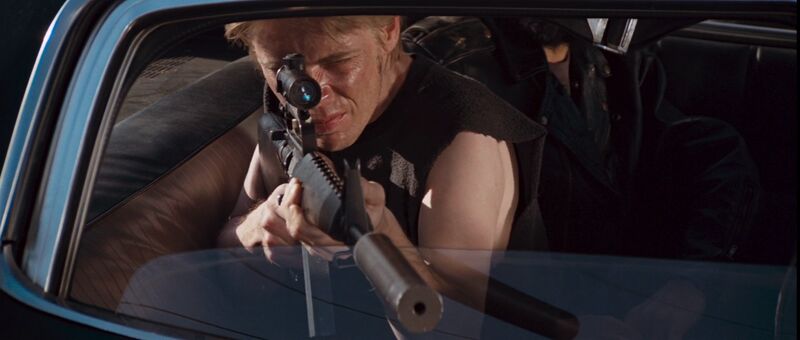 White man with blonde hair pointing a large sniper gun out of a car window