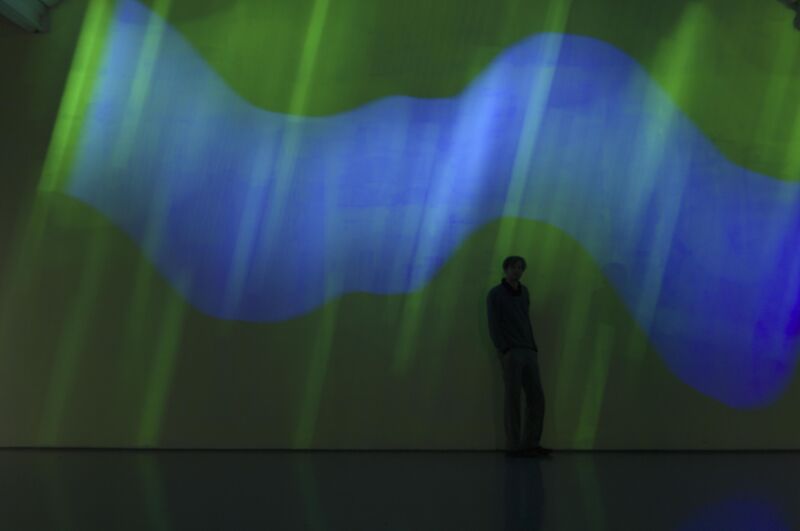 From Torsten Lauschmann's exhibition. A person walks in front of bright green and blue lights projected on DCA walls.