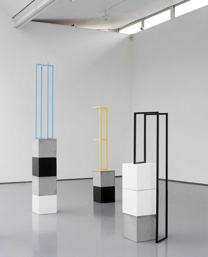 From Camilla Løw's exhibition. Black, white and grey cubes are stacked on top of each other. On top of the cubes, there are rectangular metal frames in yellow, blue and black.