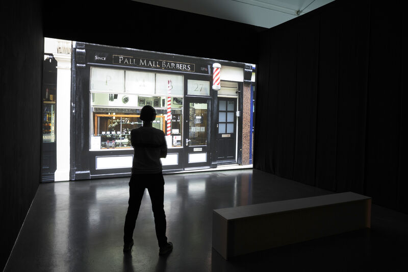 A film is projected in a dark room, while a person watches. They are standing, there is also a low bench to the right of the photograph. The film still shows a pub front. 