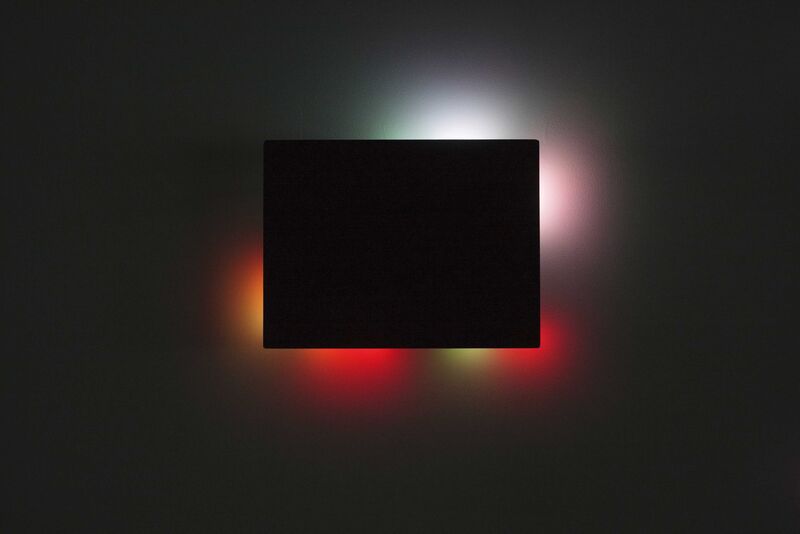 From Jim Campbell's exhibition. A black rectangular canvas blocks multi-coloured lights, but some light from the lights can still be seen from behind it.