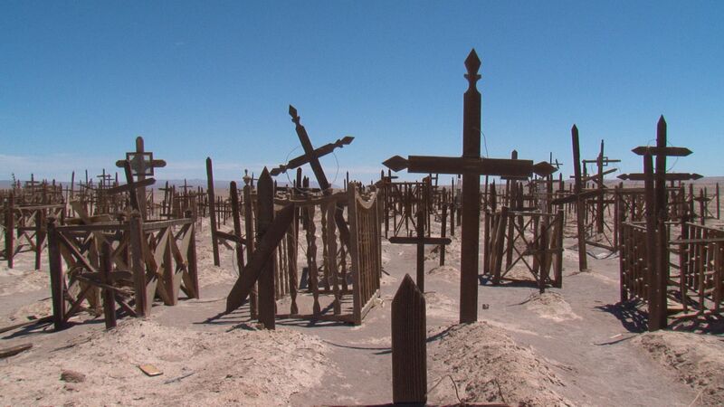 A collection of wooden crosses stand in a desert with a bright blue sky behind.
