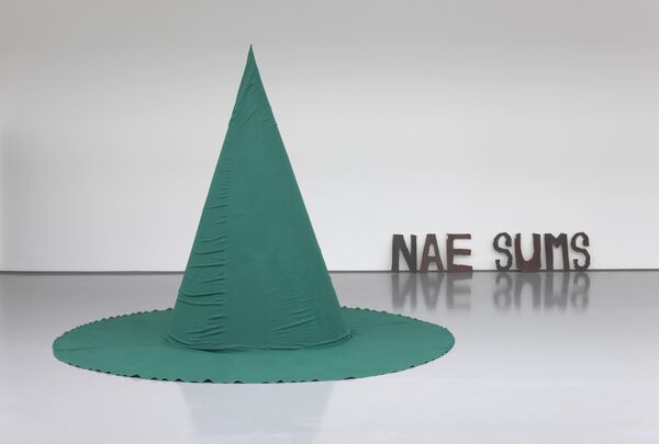 From Ruth Ewan's exhibition. A teal-green object shaped like a witches' hat sits on the floor. Behind, there are the words 'Nae Sums'
