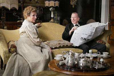 Two people in period clothing sit on a yellow upholstered sofa in an ornate room, with one person holding a large newspaper and a silver tea service set on a wooden table in the foreground.