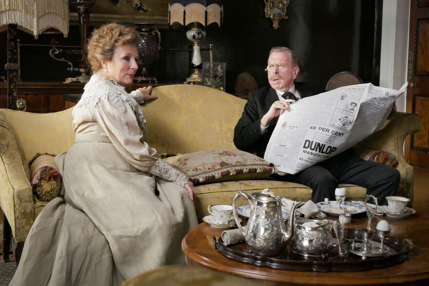 Two people in period clothing sit on a yellow upholstered sofa in an ornate room, with one person holding a large newspaper and a silver tea service set on a wooden table in the foreground.