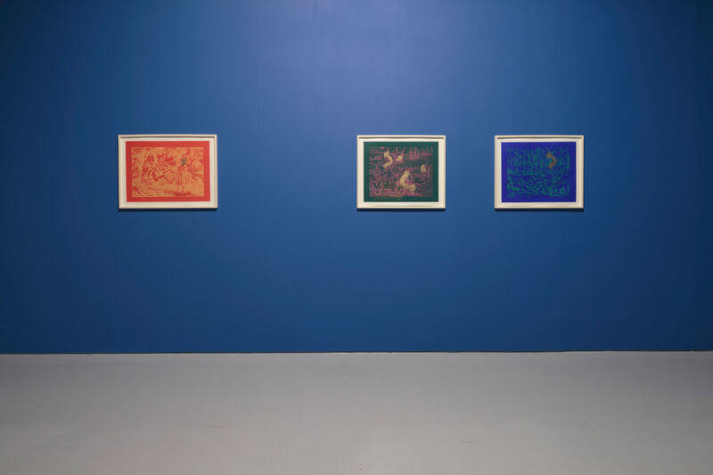 Three framed works on a bright blue wall. Left to right, these are mainly red, green, and blue, and they all have a white border.  