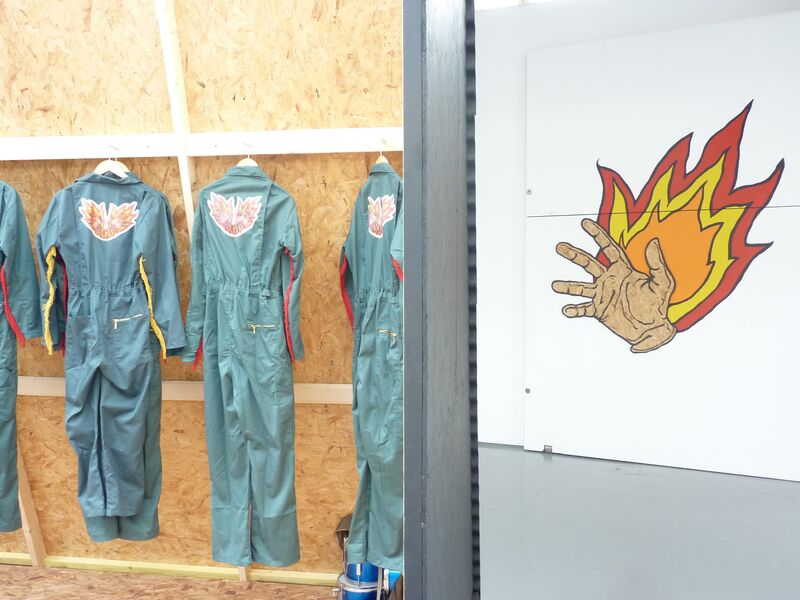 Boiling suits hang on hangers. They are teal and have a logo of a hand with flames coming out of it on the back.