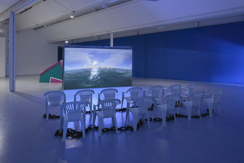 An installation image of gallery two showing a green structure like a building with a corrugated roof and a pink trim, as though it is sinking into the floor. On the back of one of these structures a film is projected. White plastic chairs are positioned in front of the screen, with iron chains around their bases. The film still shows the sea.