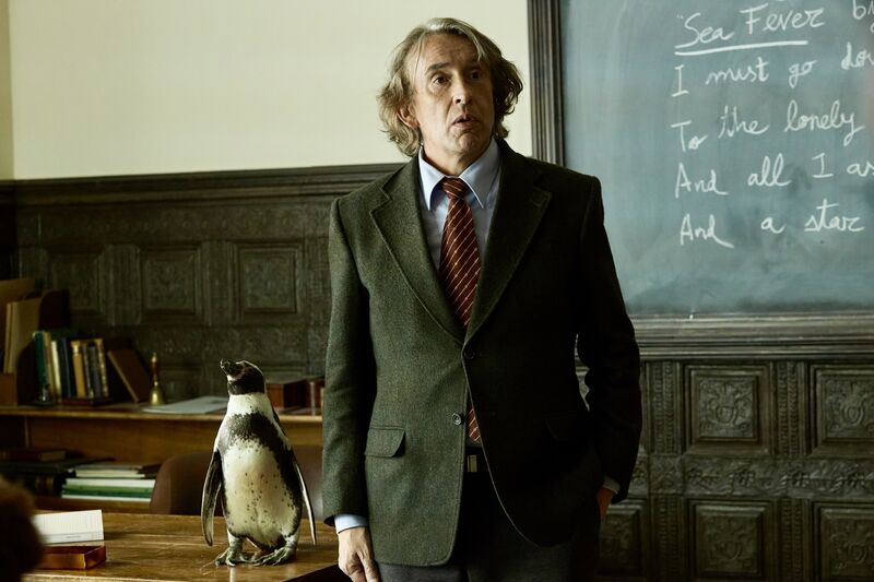 A man in a suit stands in a classroom next to a penguin.