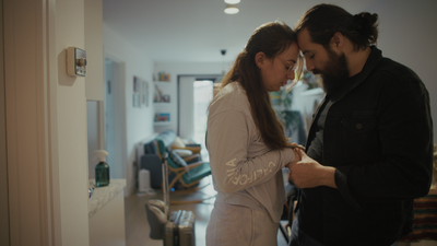 Two people stand close together in a modern apartment hallway, with one person holding the other's hands, while a living area with chairs, shelves and soft natural light is visible in the background.