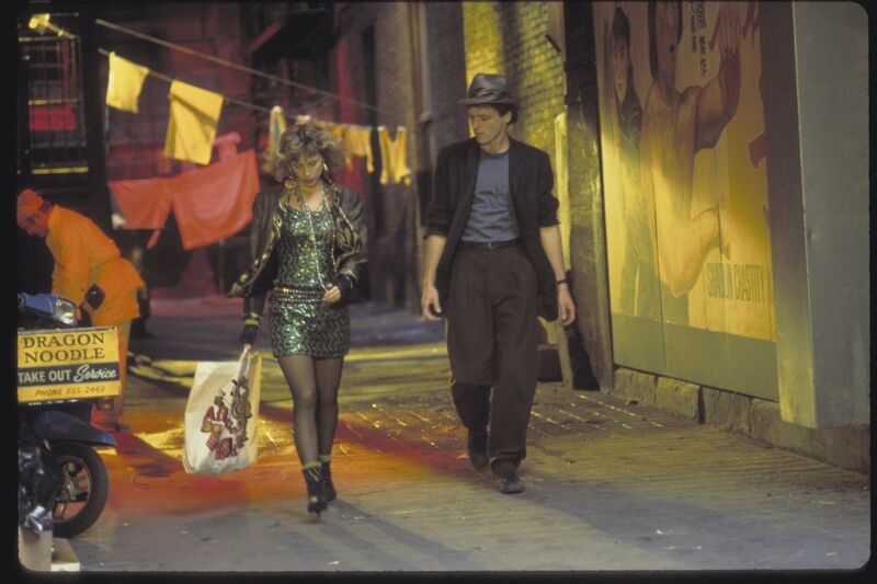 A man and a woman, both wearing eighties fashion, walk through a busy city street together.
