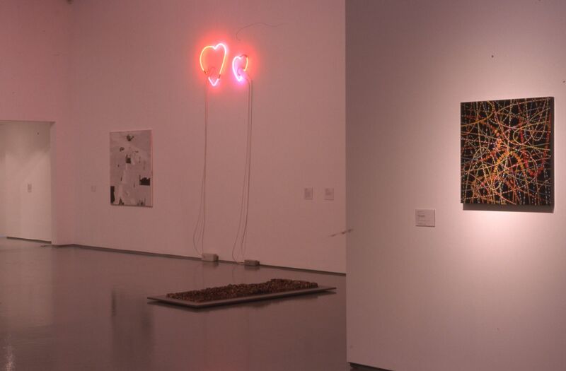 A pink, holographic light hangs from DCA Galleries in the shape of a heart. A black, abstract painting hangs next to this.